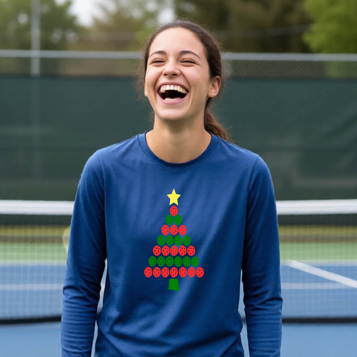 A woman laughing on a pickleball  court wearing a blue long-sleeve shirt with a Christmas tree made of red and green tpickleballs and a yellow star.