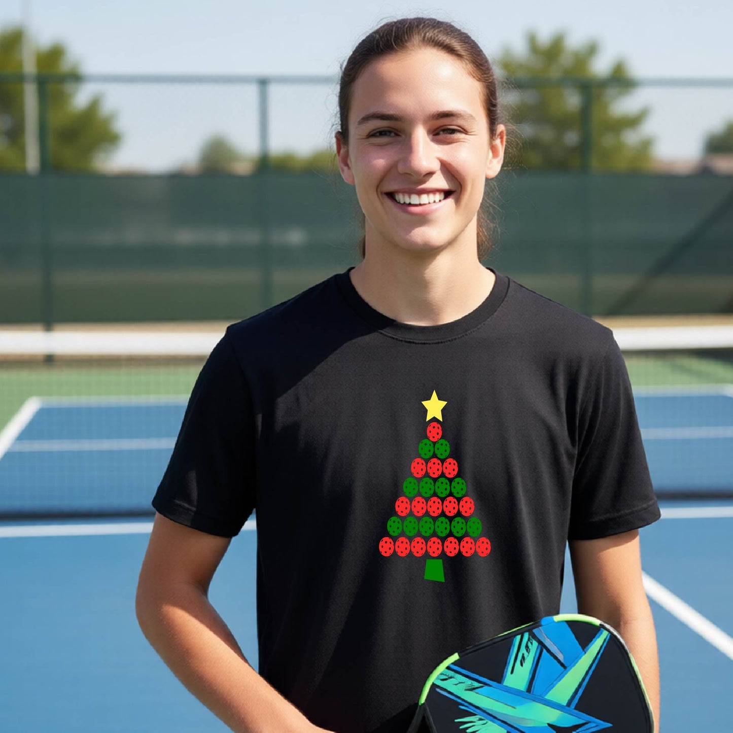A smiling woman on a blue pickleball court wearing a black t-shirt with a Christmas tree made of red and green pickleballs and a yellow star, holding a pickleball paddle.