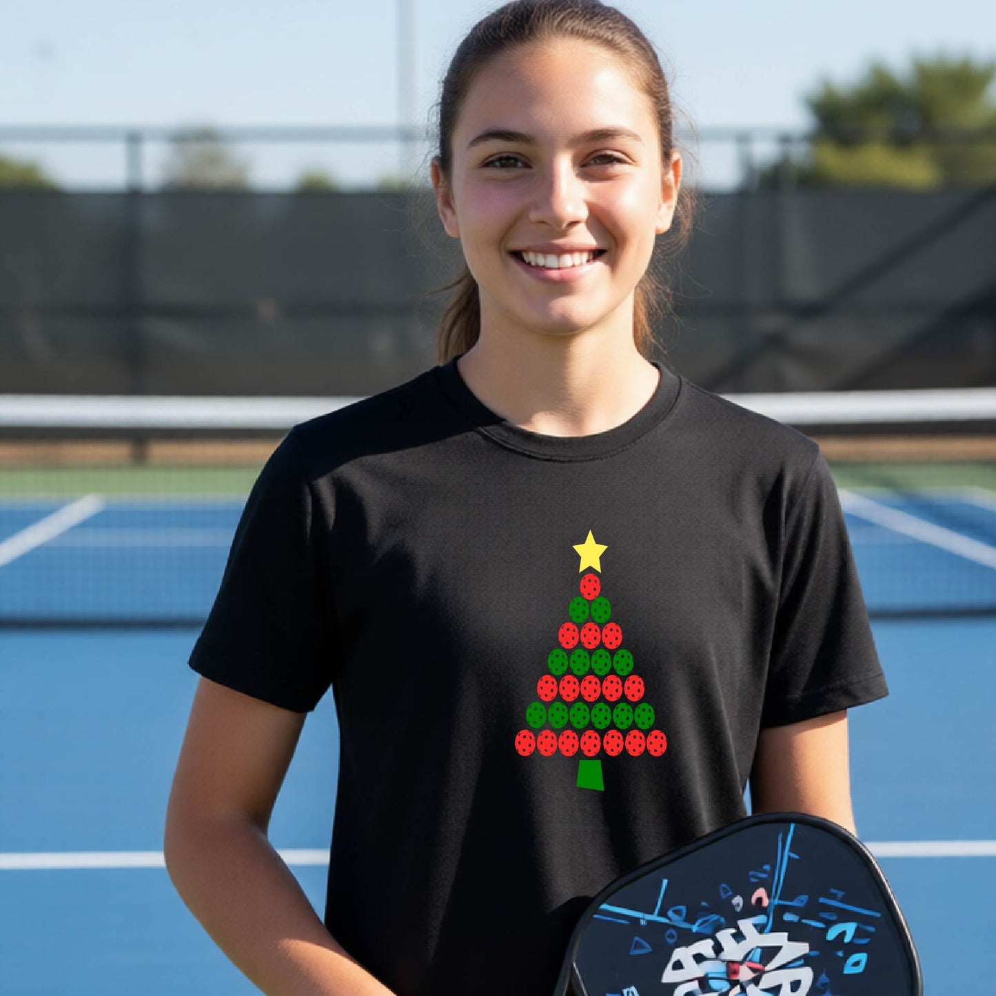 A smiling young woman on a blue pickleball court wearing a black t-shirt with a Christmas tree made of red and green pickleball and a yellow star. She is holding a pickleball paddle.