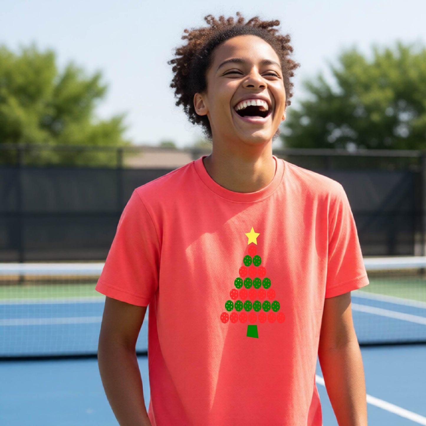 A smiling person standing on a blue pickleball  court wearing a coral-pink T-shirt with a Christmas tree made of green and red pickleballs and a yellow star on the front.