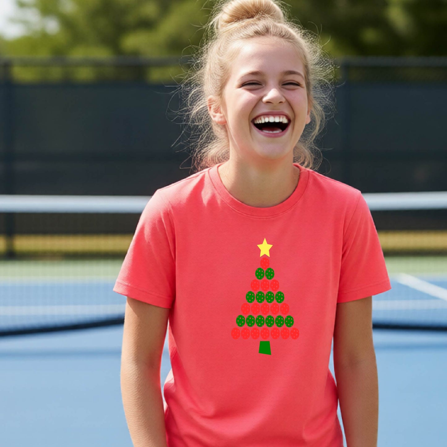 A young girl with blonde hair in a bun laughing on an outdoor pickleball court, wearing a coral pink t-shirt with a Christmas tree graphic made of green and red pickleballs and a yellow star.