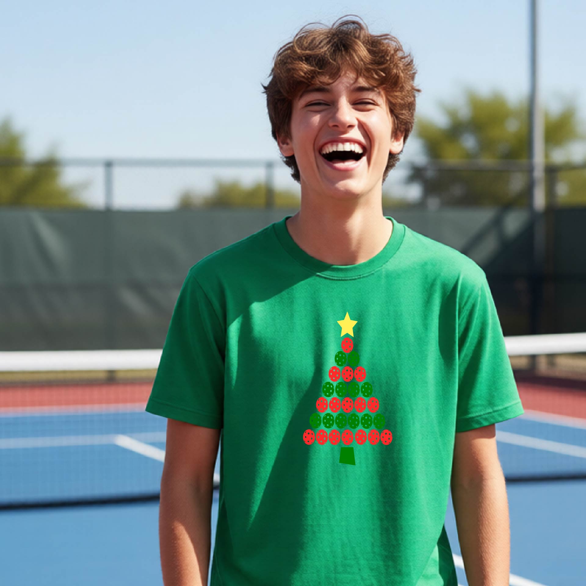 A young man laughing on an outdoor pickleball  court wearing a bright green t-shirt with a Christmas tree made of red and green pickleballs and a yellow star.