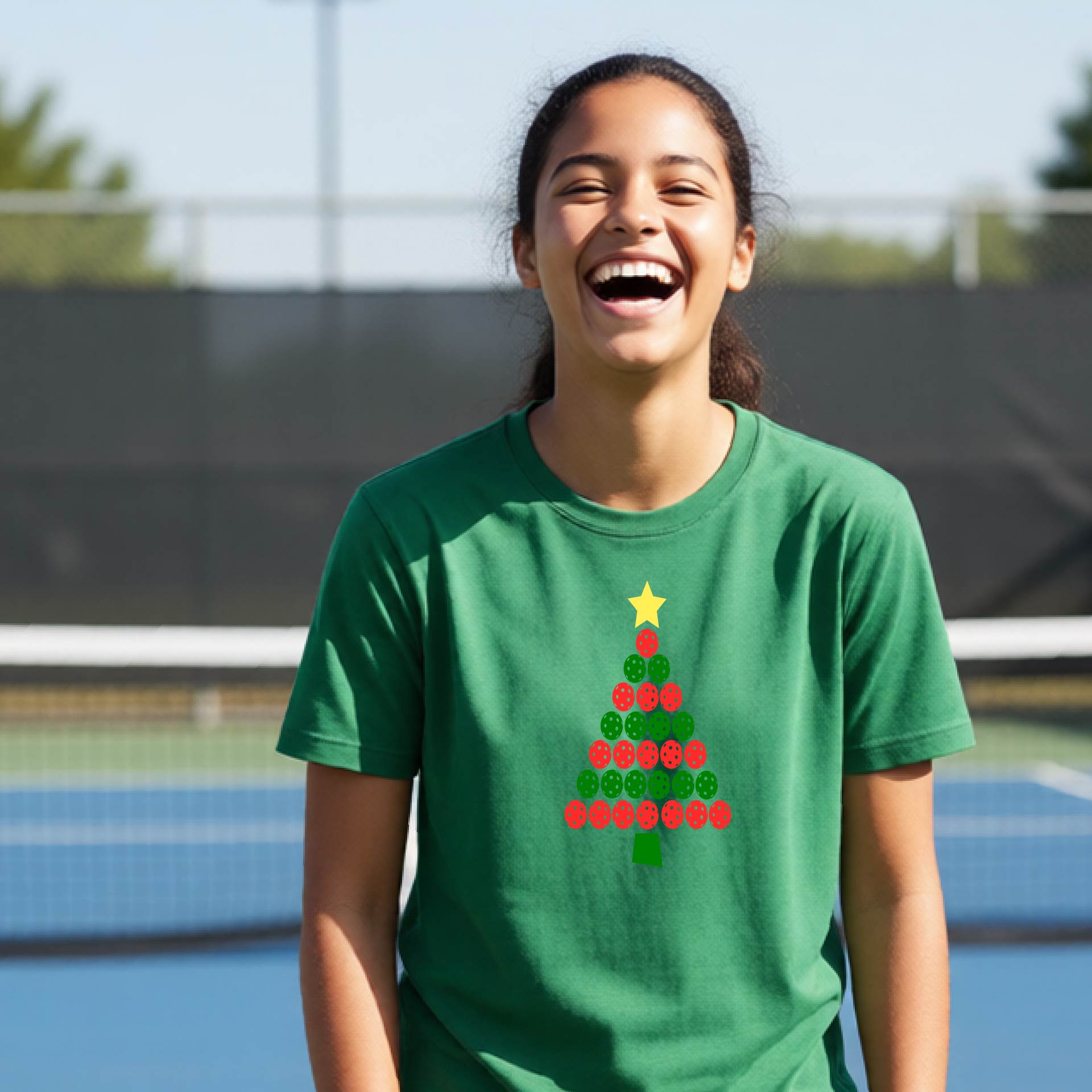 A young woman laughing on an outdoor pickleball  court wearing a green t-shirt with a Christmas tree made of red and green pickleballs and a yellow star.