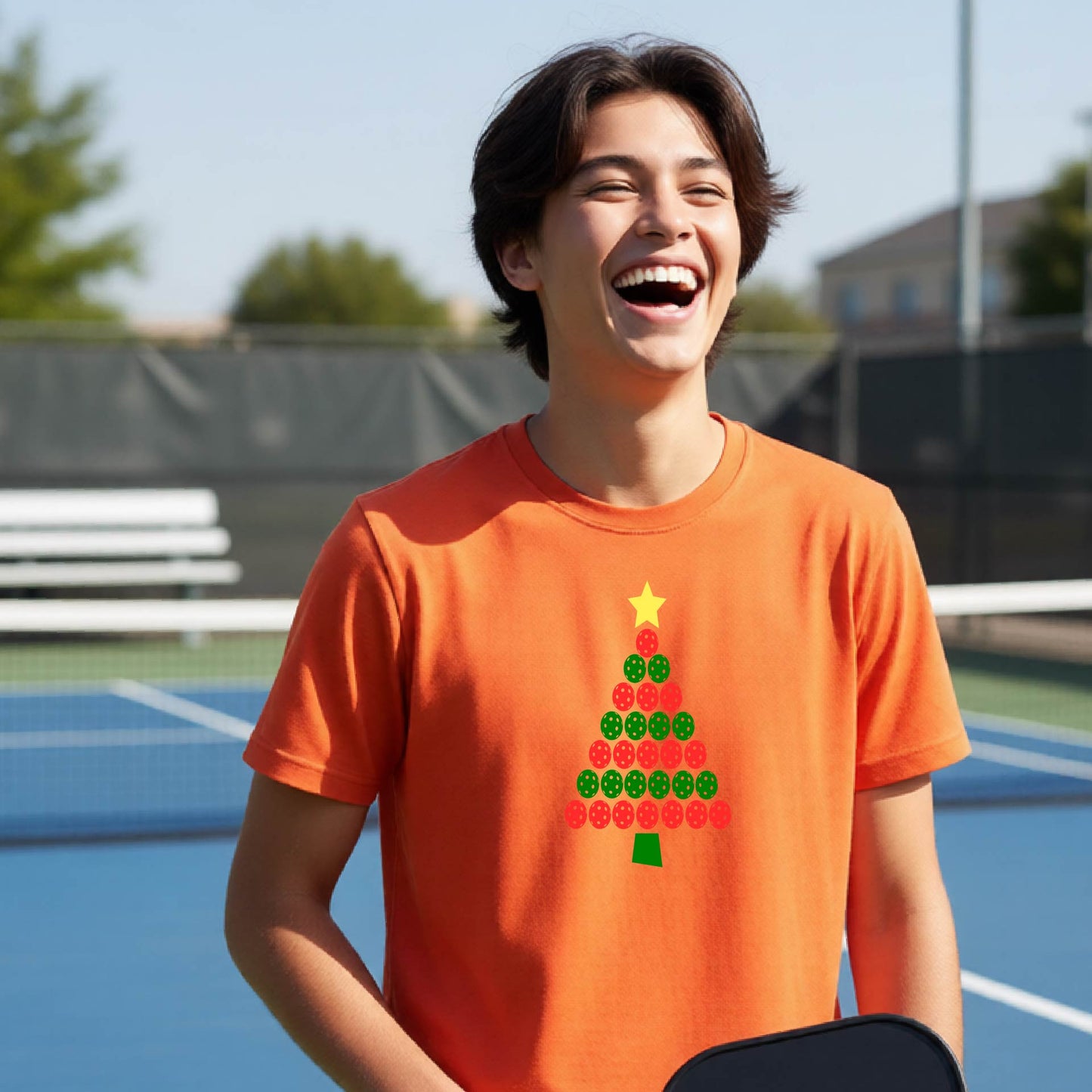 A young man laughing on an outdoor pickleball  court wearing a bright orange t-shirt with a Christmas tree made of red and green pickleballs and a yellow star.