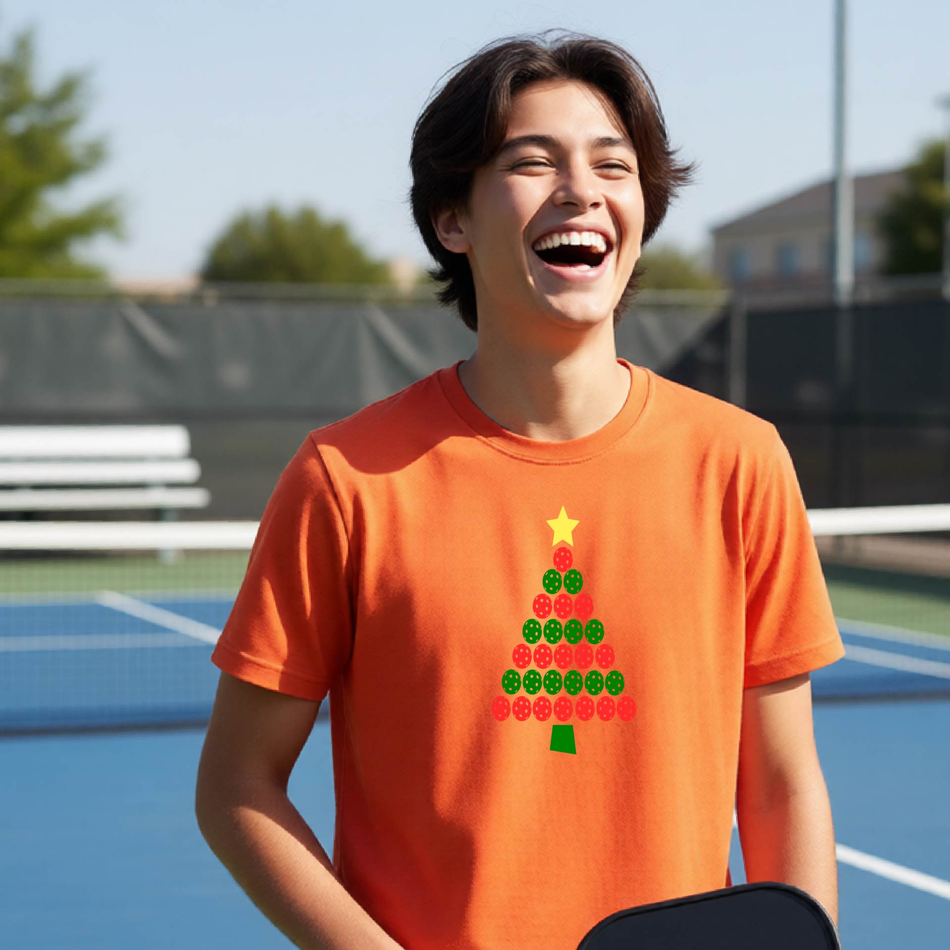 A young man laughing on an outdoor pickleball  court wearing a bright orange t-shirt with a Christmas tree made of red and green pickleballs and a yellow star.