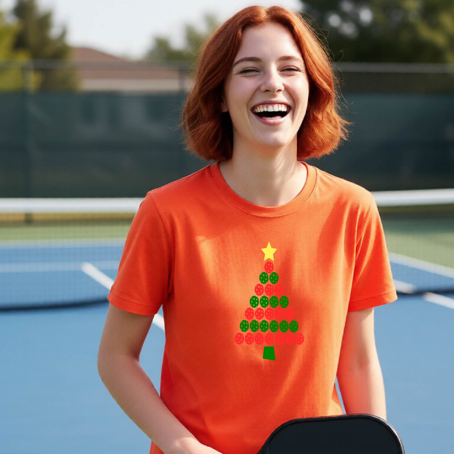 A smiling woman with red hair stands on a blue pickleball  court wearing an orange T-shirt with a Christmas tree made of green and red pickleball circular shapes and a yellow star.