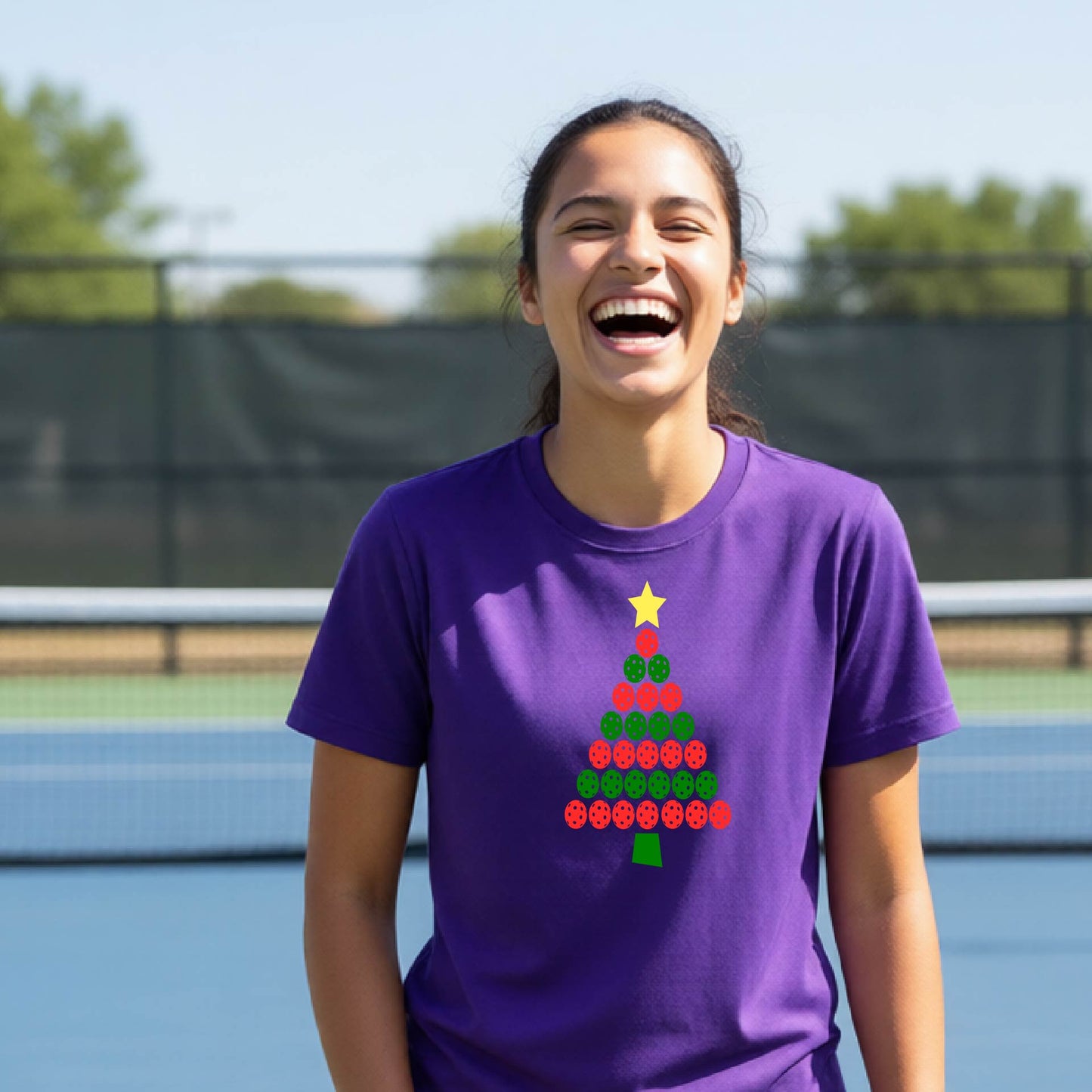 A woman laughing on an outdoor pickleball  court wearing a purple t-shirt with a Christmas tree made of pickleballs graphic.