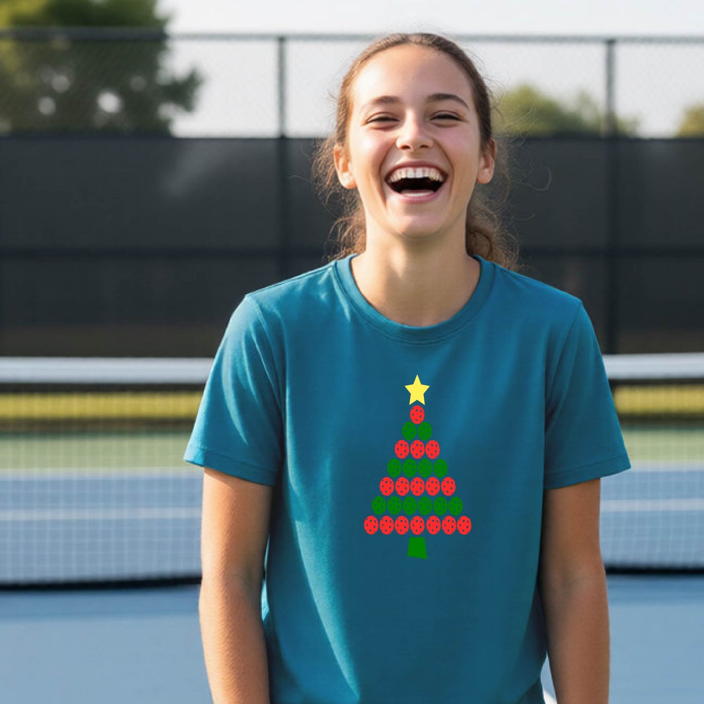 A young woman laughing on an outdoor pickleball court, wearing a teal t-shirt with a Christmas tree made of red pickleballs and a yellow star.
