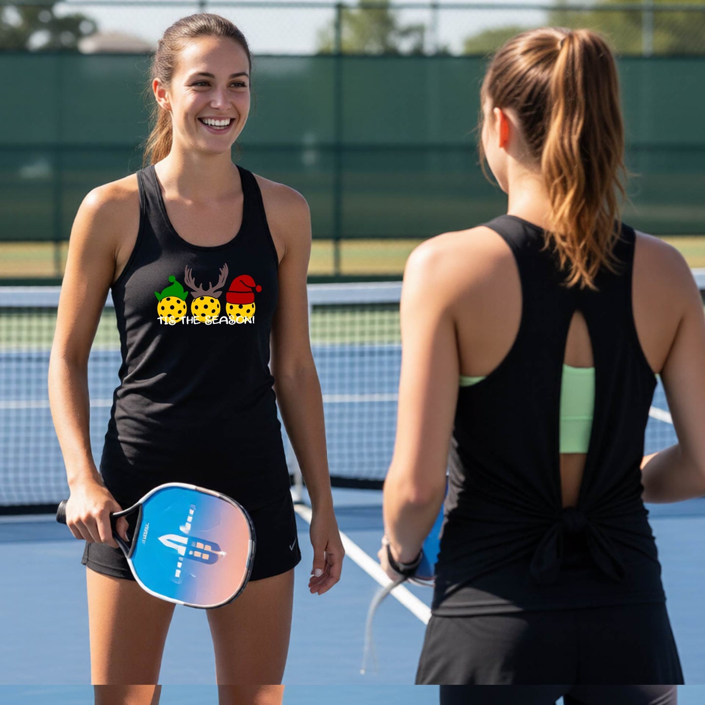 Two women wearing a black graphic split-black sleeveless tank top t-shirt with a Christmas-themed design of three pickleballs wearing holiday hats and the text 'TIS THE SEASON!'. The background shows a festive blurred Christmas tree and a pickleball court.