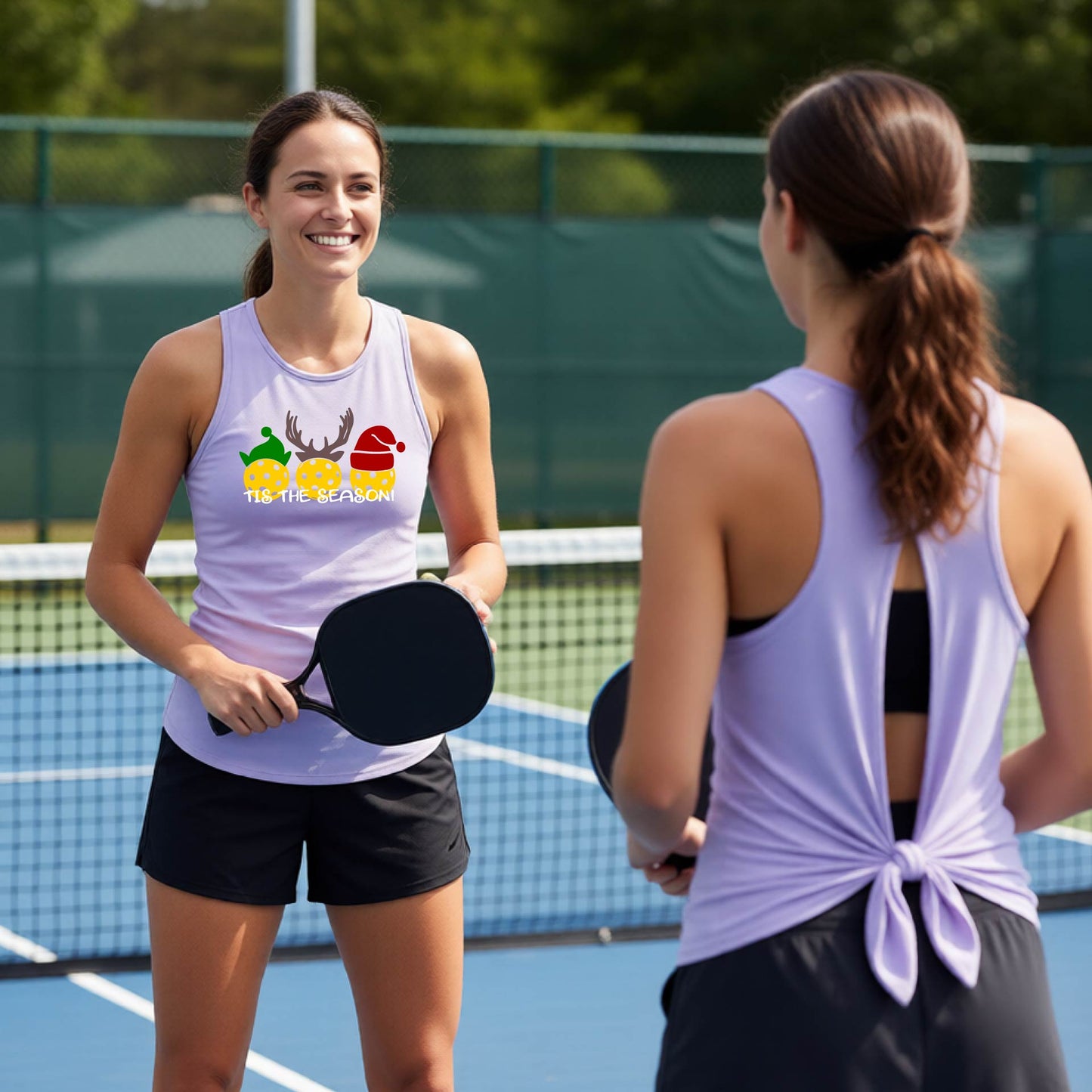 Two women wearing a pastel purple lavender graphic split-black sleeveless tank top shirt with a Christmas-themed design of three pickleballs wearing holiday hats and the text 'TIS THE SEASON!'. The background shows a festive blurred Christmas tree and a pickleball court.