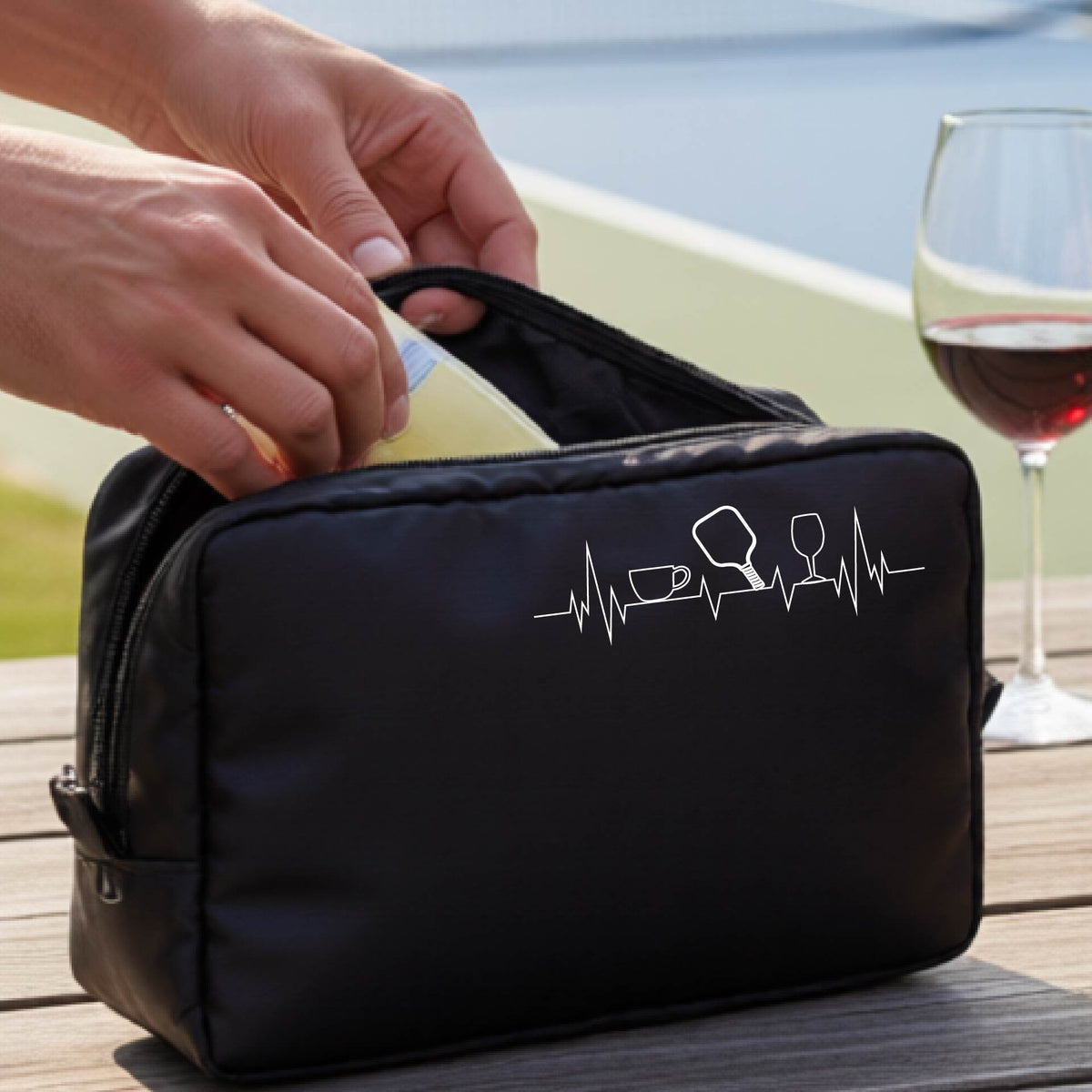 A person holds a black zippered cosmetic pouch outdoors on a wooden table, with a pickleball court blurred in the background. The pouch has a white line-art graphic of a coffee cup, pickleball paddle and a wine glass, on a heartbeat line.