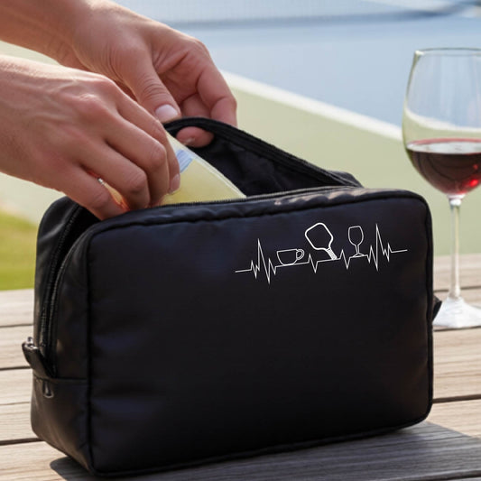 A person holds a black zippered cosmetic pouch outdoors on a wooden table, with a pickleball court blurred in the background. The pouch has a white line-art graphic of a coffee cup, pickleball paddle and a wine glass, on a heartbeat line.