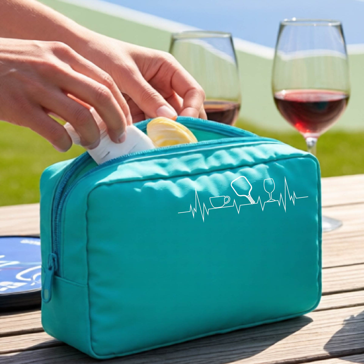 A person holds an aqua blue zippered cosmetic pouch outdoors on a wooden table, with a pickleball court blurred in the background. The pouch has a white line-art graphic of a coffee cup, pickleball paddle and a wine glass, on a heartbeat line.