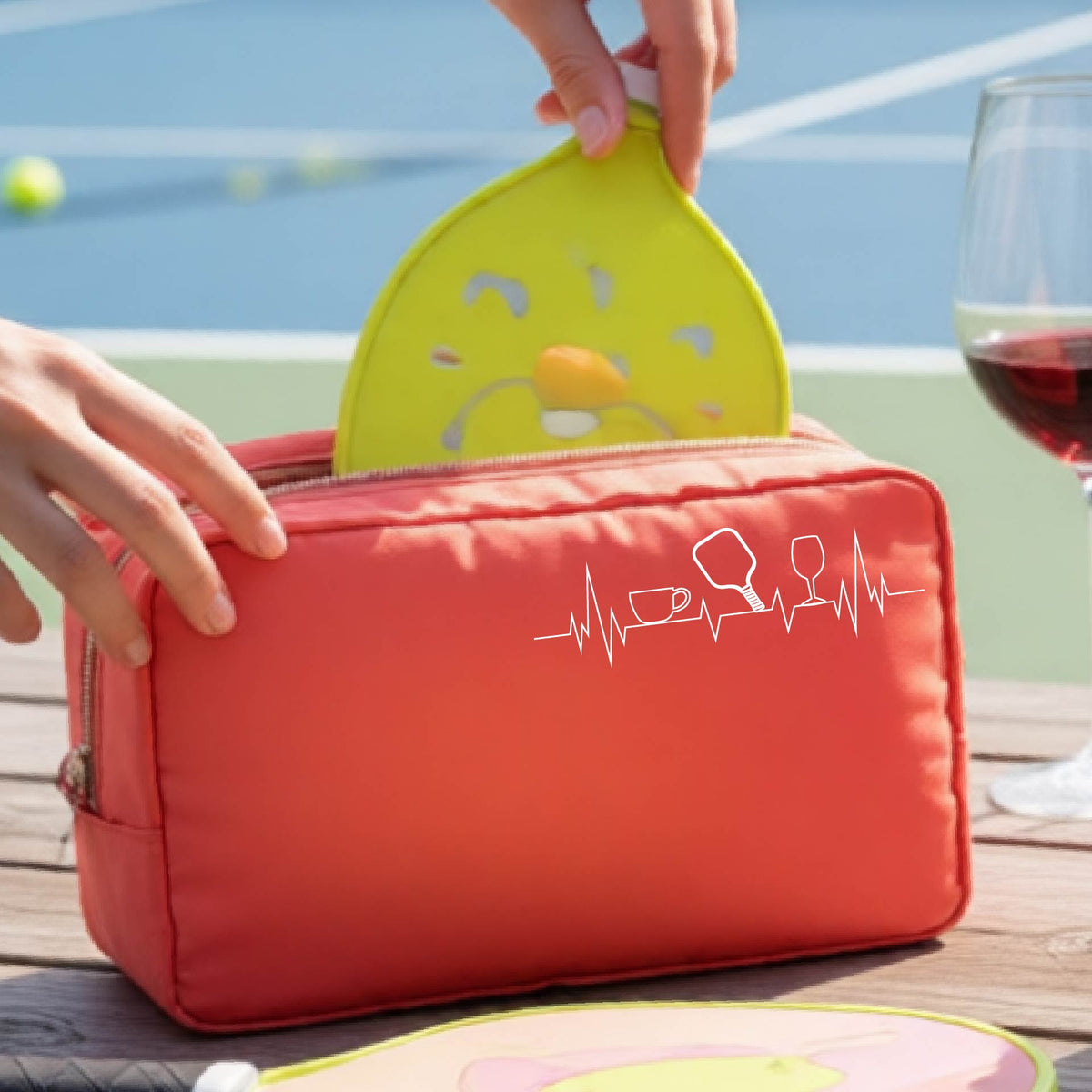 A person holds a coral-colored zippered cosmetic pouch outdoors on a wooden table, with a pickleball court blurred in the background. The pouch has a white line-art graphic of a coffee cup, pickleball paddle and a wine glass, on a heartbeat line.