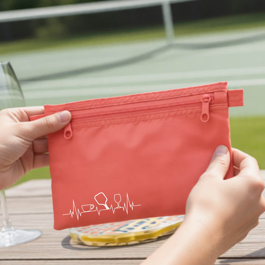 A person holds a coral pink zippered cosmetic pouch outdoors on a wooden table, with a pickleall court blurred in the background. The pouch has a white line-art graphic of a coffee cup, pickleball paddle and a wine glass, on a heartbeat line. 