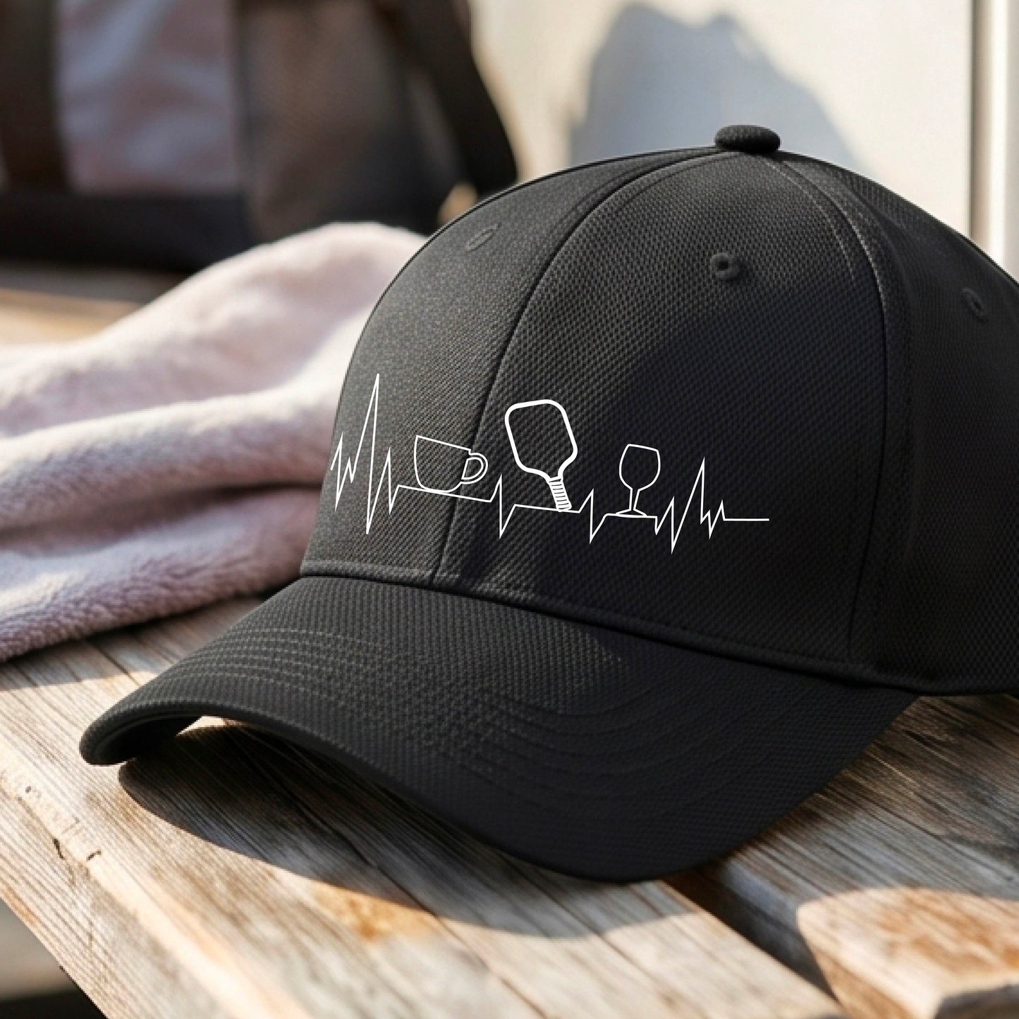 A close up of a black pickleball visor with  a white line-art graphic of a coffee cup, pickleball paddle and a wine glass, on a heartbeat EKG line.