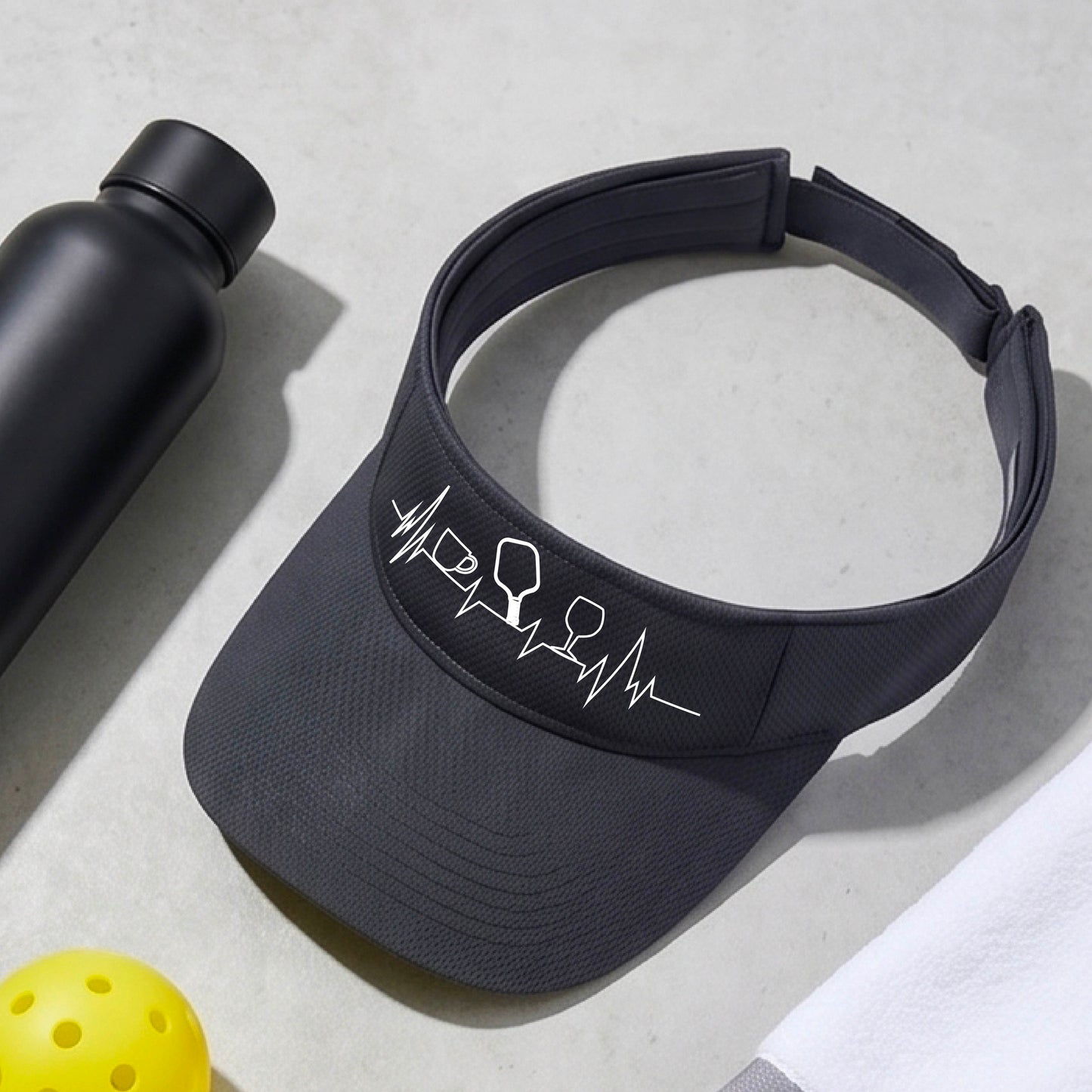 A close up of a navy blue pickleball visor with  a white line-art graphic of a coffee cup, pickleball paddle and a wine glass, on a heartbeat EKG line.