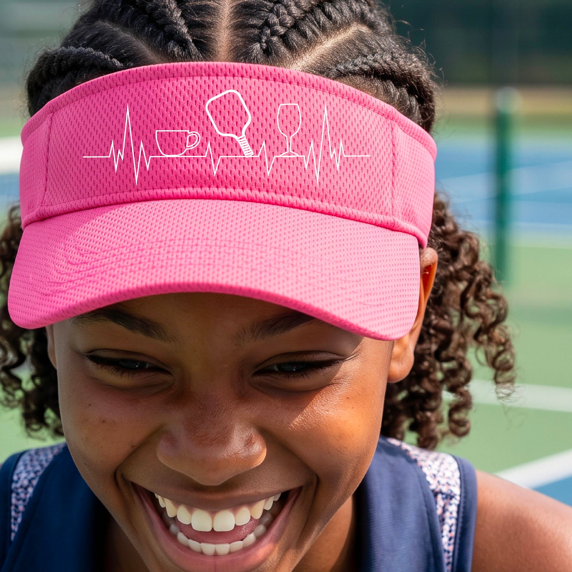 A close up of a pink pickleball visor with  a white line-art graphic of a coffee cup, pickleball paddle and a wine glass, on a heartbeat EKG line.