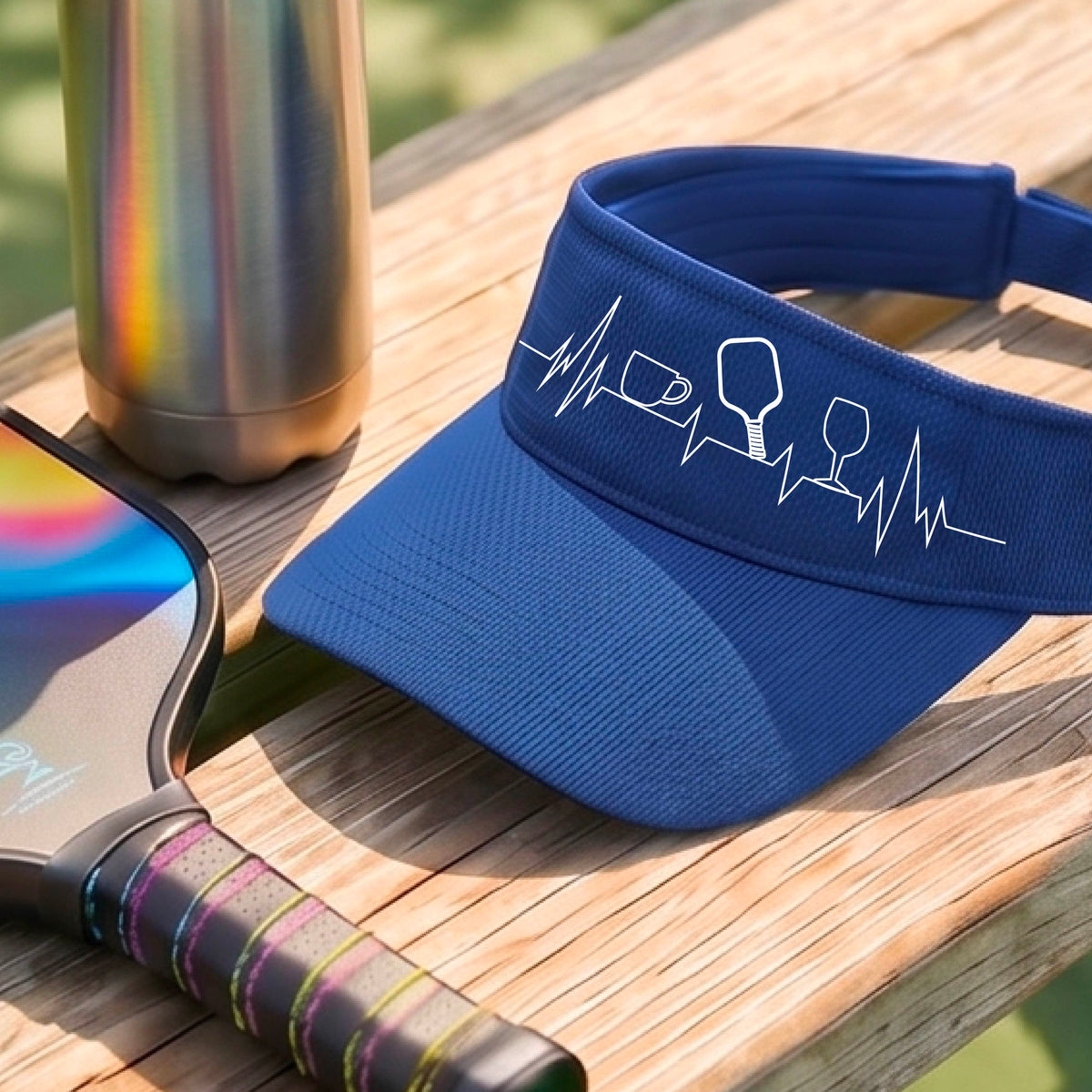 A close up of a royal blue pickleball visor with  a white line-art graphic of a coffee cup, pickleball paddle and a wine glass, on a heartbeat EKG line.