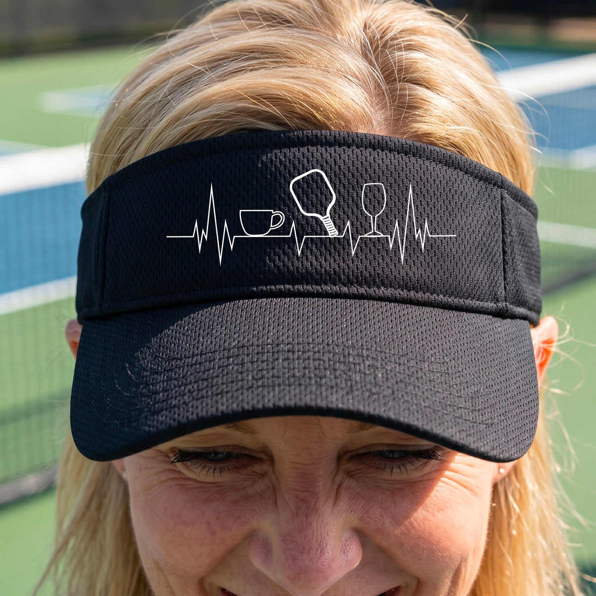 A close up of a black pickleball visor with  a white line-art graphic of a coffee cup, pickleball paddle and a wine glass, on a heartbeat EKG line.