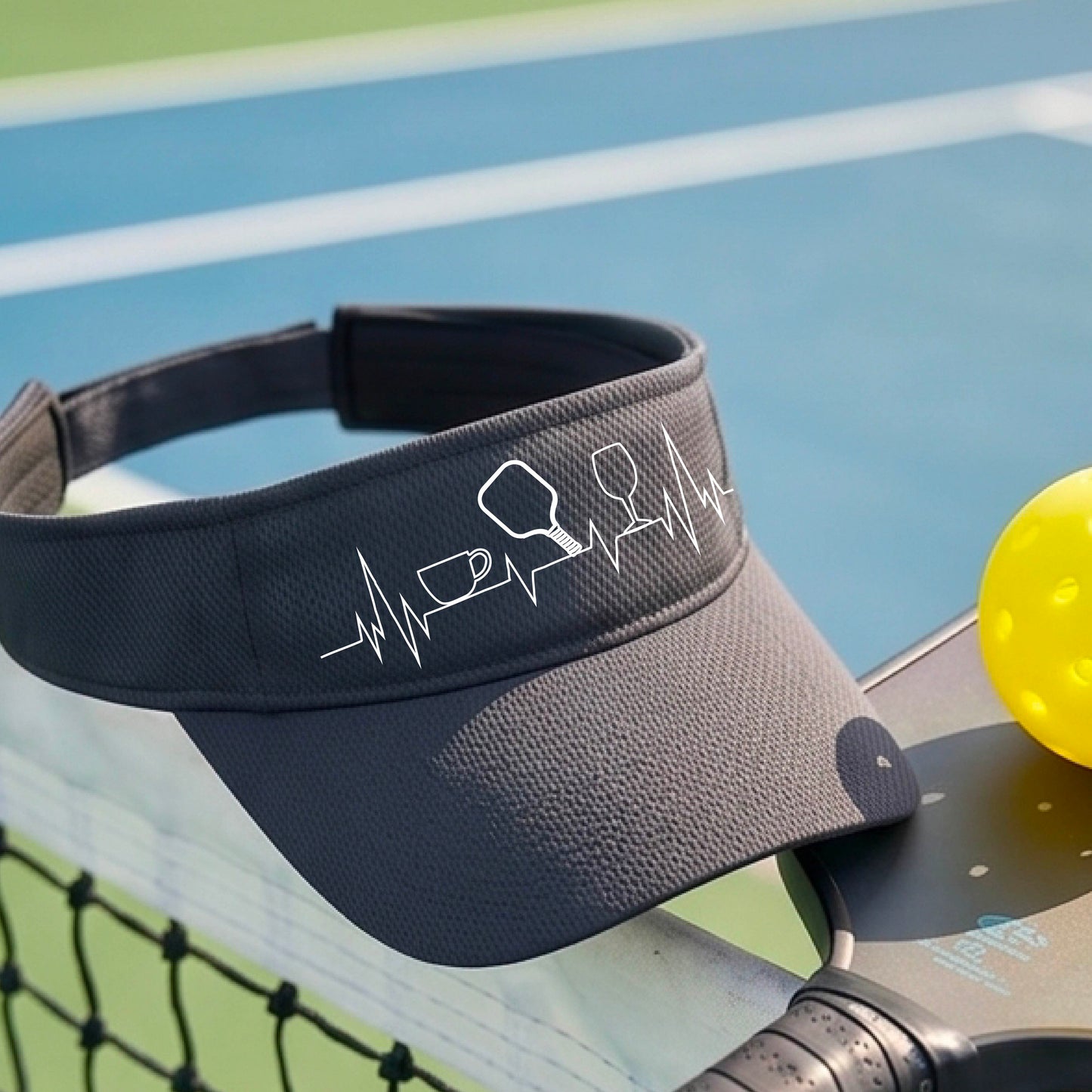 A close up of a gray pickleball visor with  a white line-art graphic of a coffee cup, pickleball paddle and a wine glass, on a heartbeat EKG line.