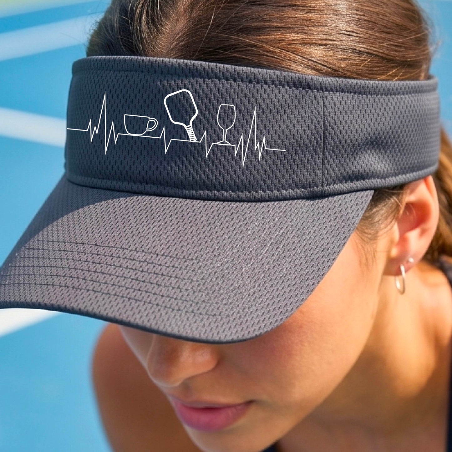 A close up of a gray pickleball visor with  a white line-art graphic of a coffee cup, pickleball paddle and a wine glass, on a heartbeat EKG line.