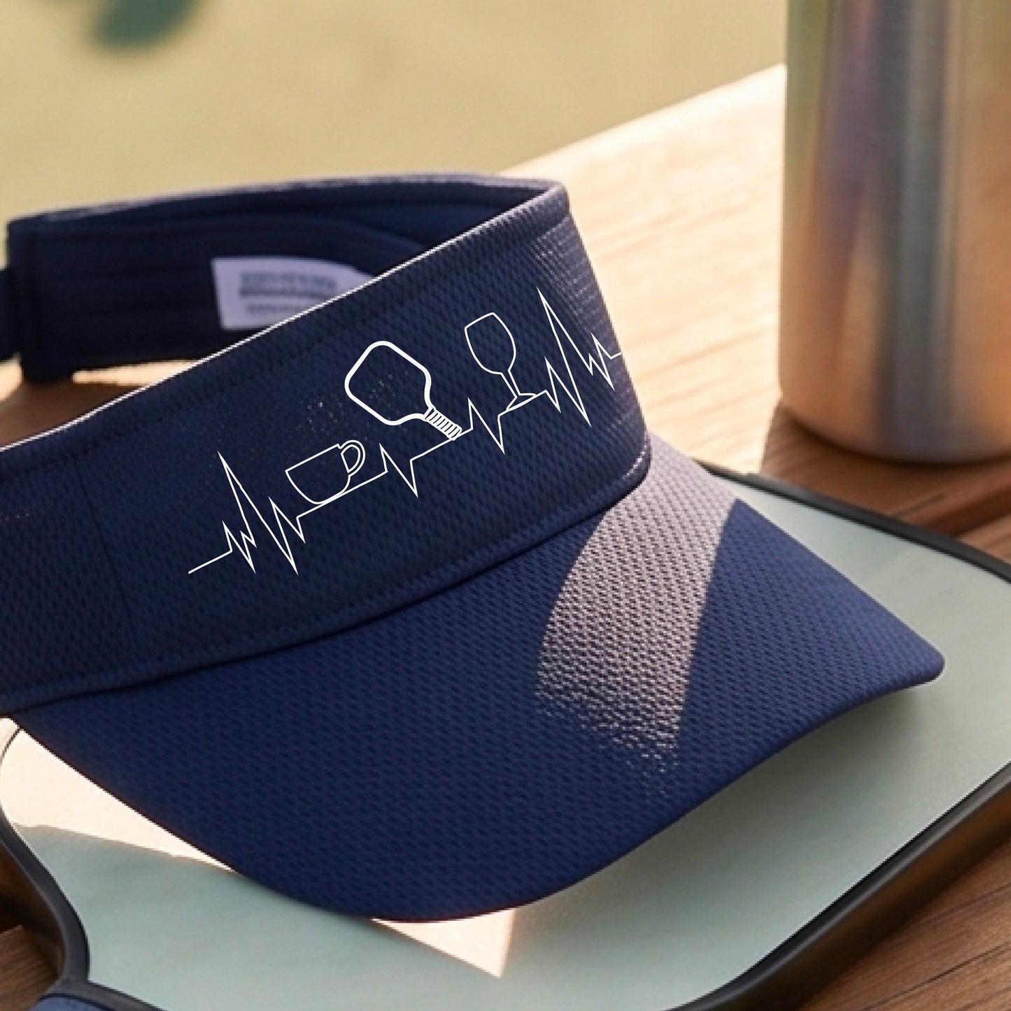 A close up of a navy blue pickleball visor with  a white line-art graphic of a coffee cup, pickleball paddle and a wine glass, on a heartbeat EKG line.