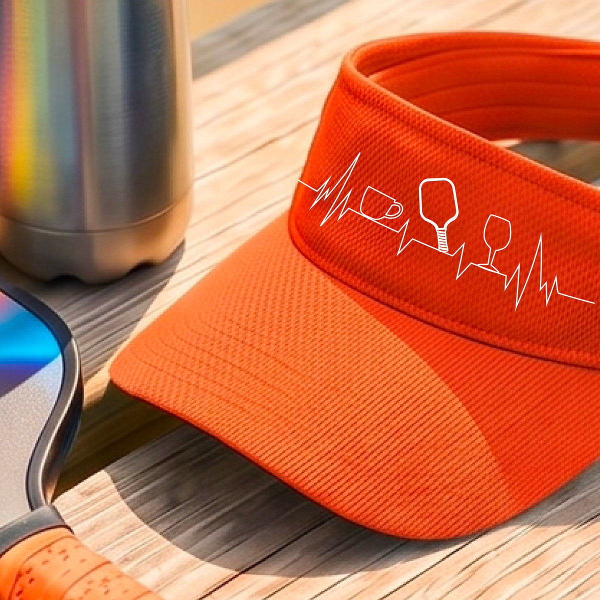 A close up of an orange pickleball visor with  a white line-art graphic of a coffee cup, pickleball paddle and a wine glass, on a heartbeat EKG line.