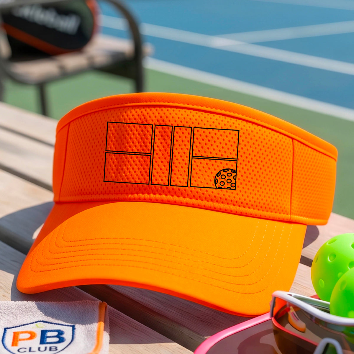 A close up of an orange visor with a black pickleball court and ball graphic on the front and a pickleball court in the background. 