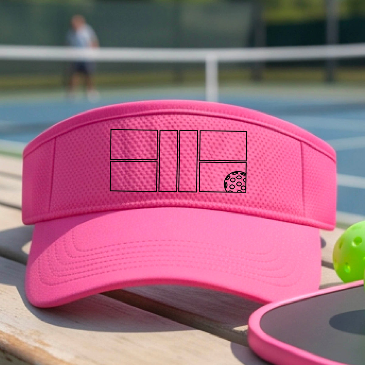 A close up of a pink visor with a black pickleball court and ball graphic on the front and a pickleball court in the background. 