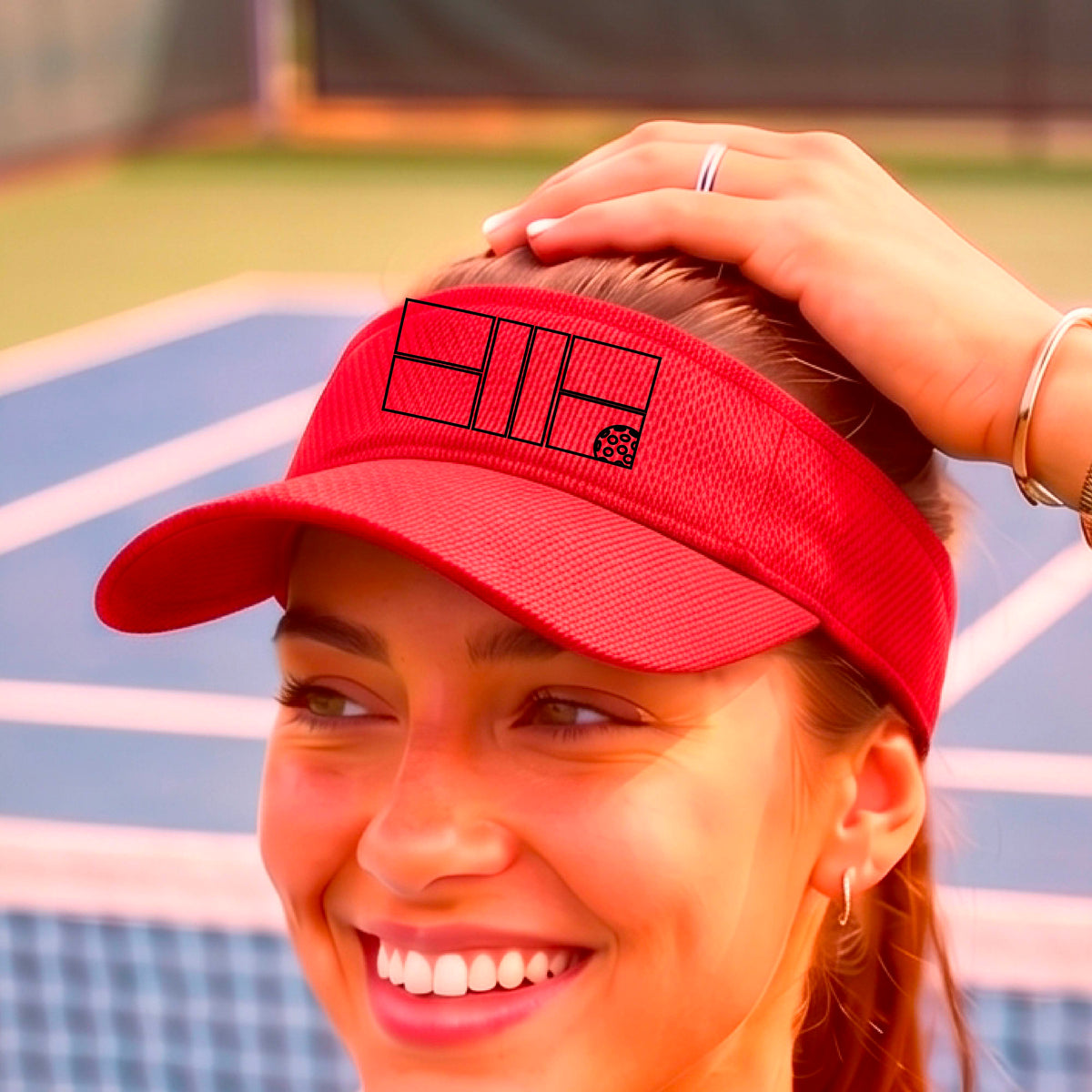 A close up of a red visors with a black pickleball court and ball graphic on the front and a pickleball court in the background. 
