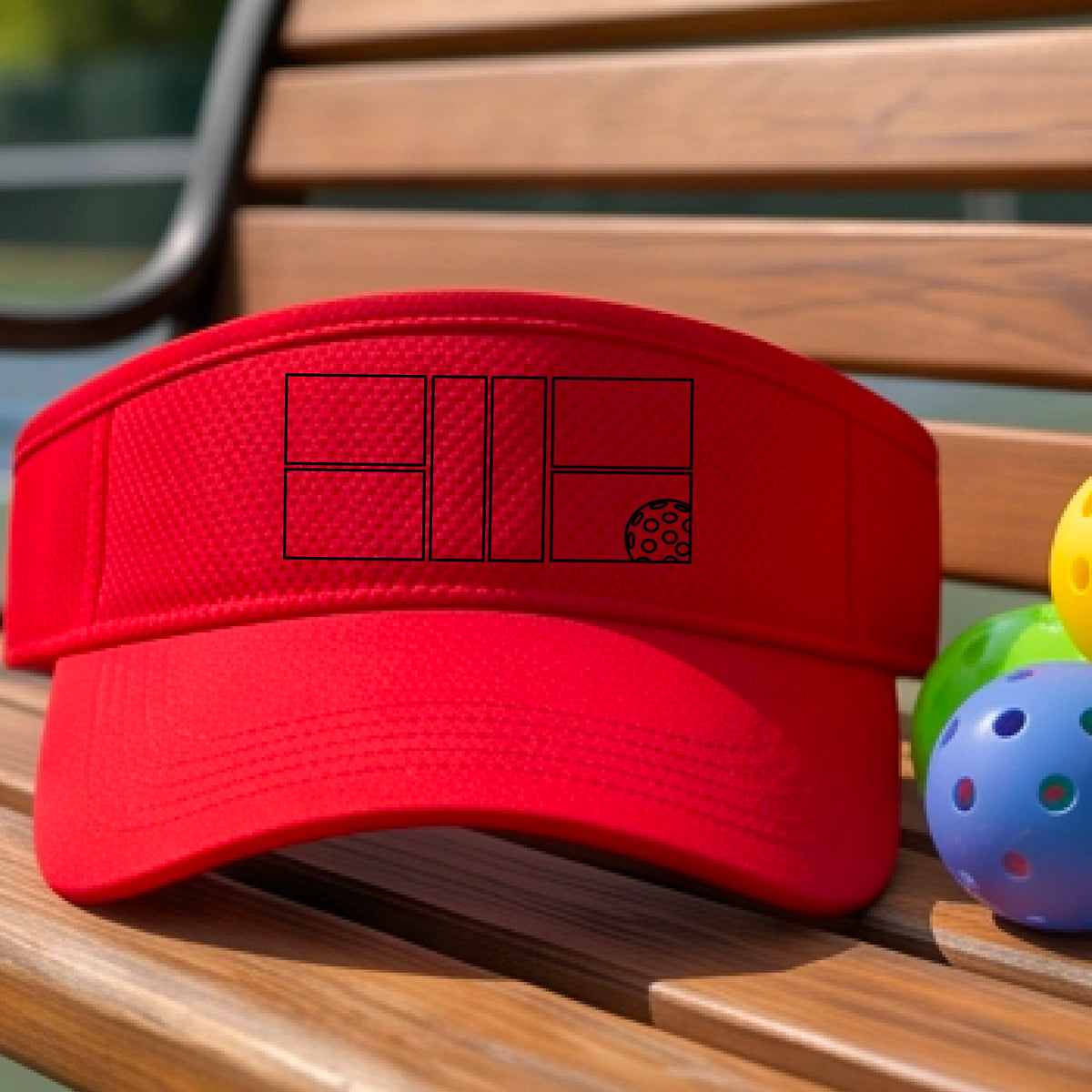 A close up of a red visor with a black pickleball court and ball graphic on the front and a pickleball court in the background. 