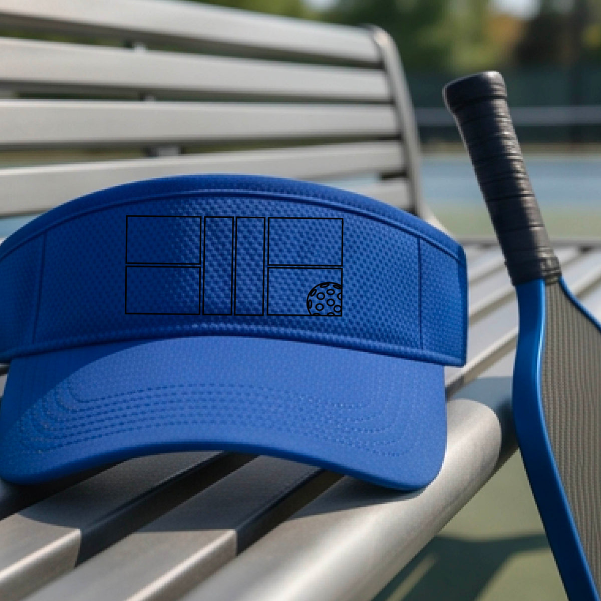 A close up of a royal  bluevisor with a black pickleball court and ball graphic on the front and a pickleball court in the background. 