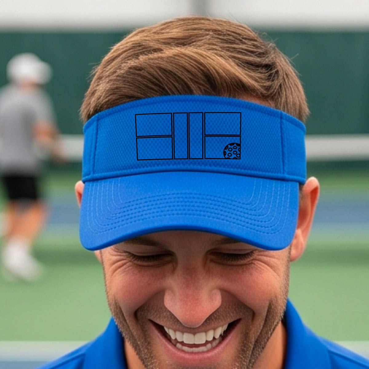 A close up of a royal blue visor with a black pickleball court and ball graphic on the front and a pickleball court in the background. 