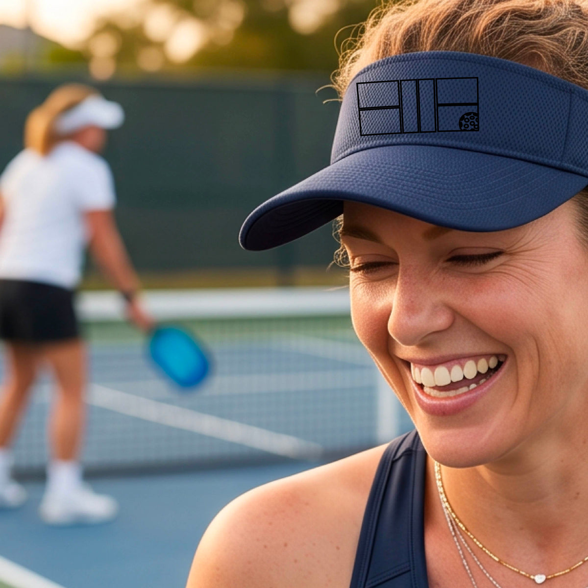 A close up of a navy blue visor with a black pickleball court and ball graphic on the front and a pickleball court in the background. 
