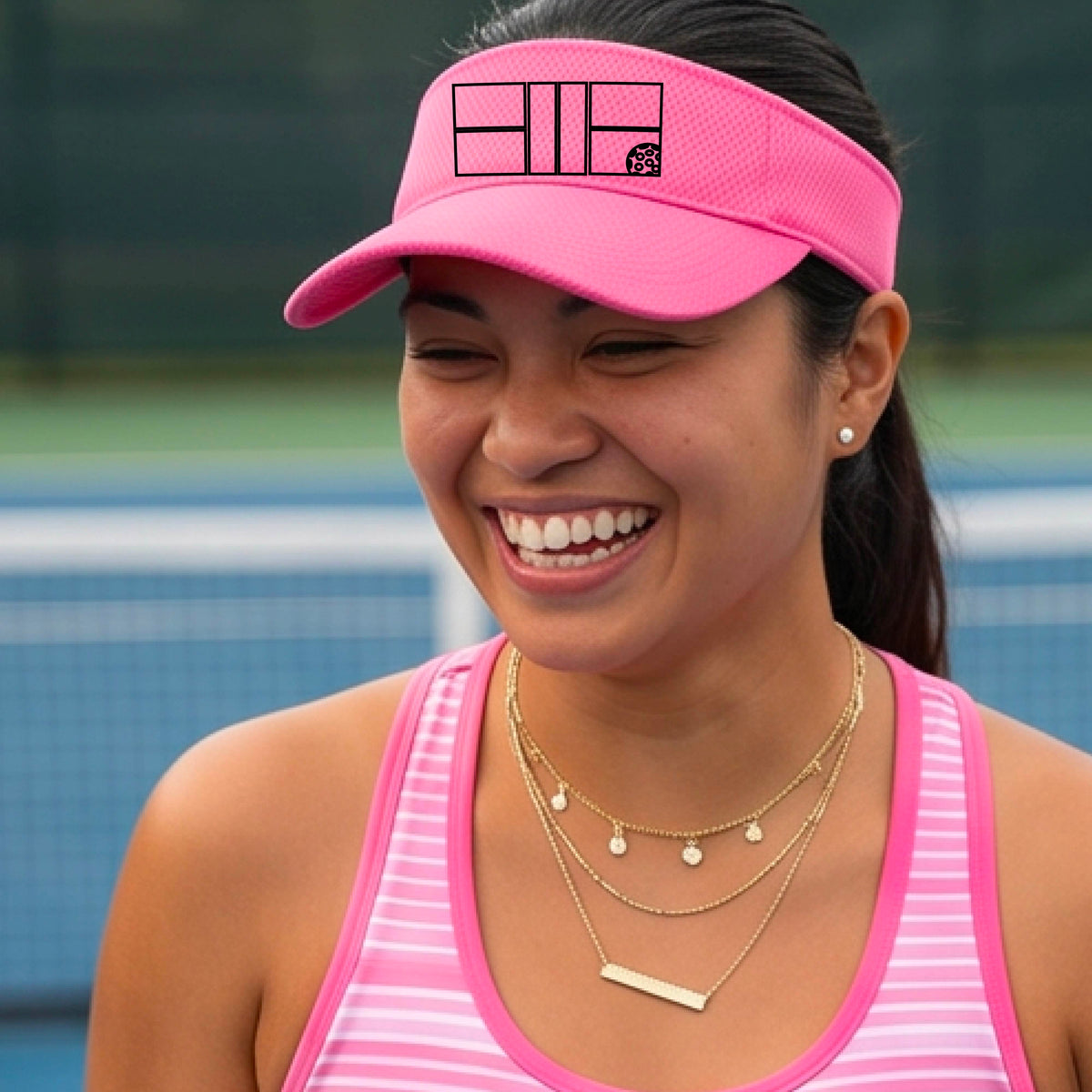A close up of a pink visor with a black pickleball court and ball graphic on the front and a pickleball court in the background. 