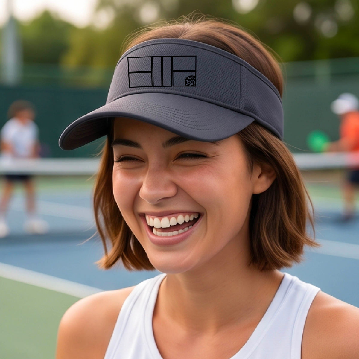 A close up of a dark gray visor with a black pickleball court and ball graphic on the front and a pickleball court in the background. 