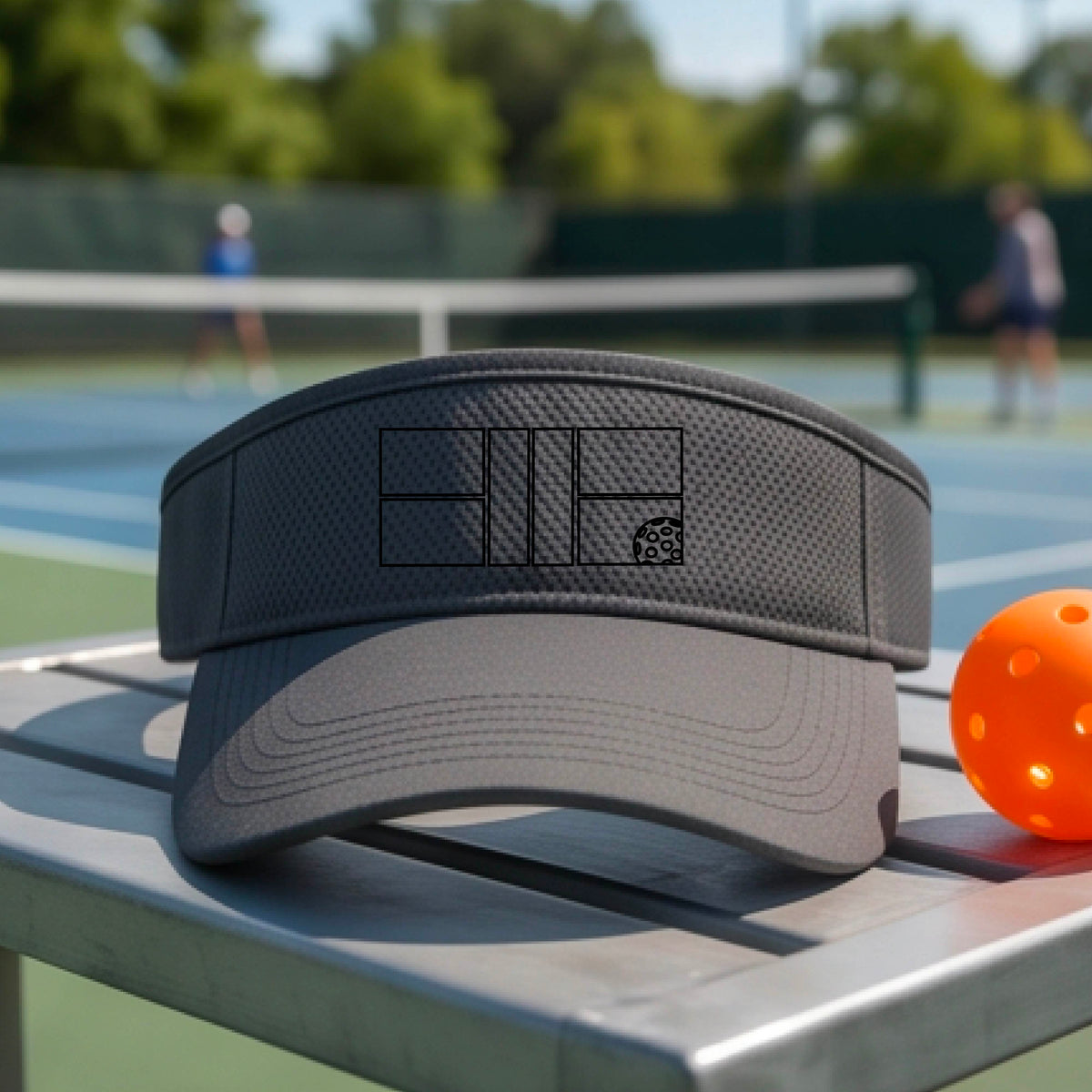 A close up of a dark gray visor with a black pickleball court and ball graphic on the front and a pickleball court in the background. 