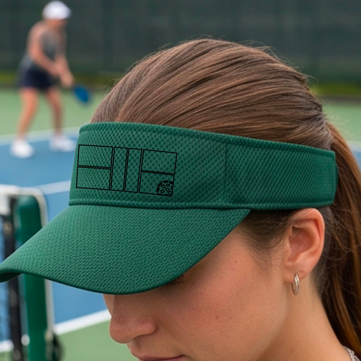 A close up of a green visor with a black pickleball court and ball graphic on the front and a pickleball court in the background. 