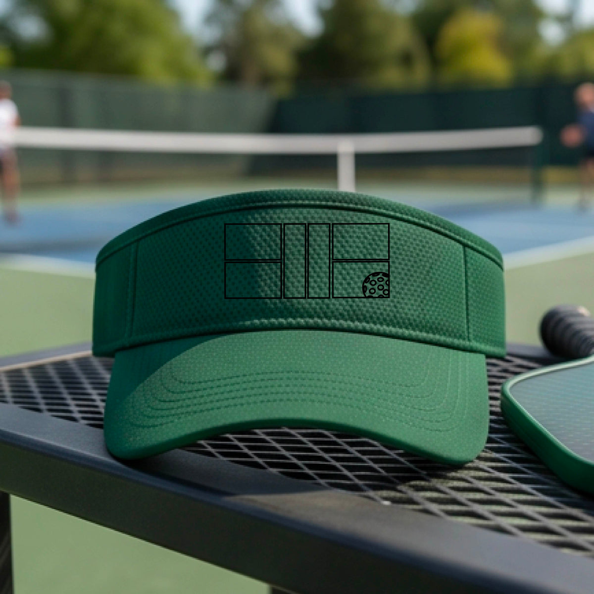 A close up of a green visor with a black pickleball court and ball graphic on the front and a pickleball court in the background. 