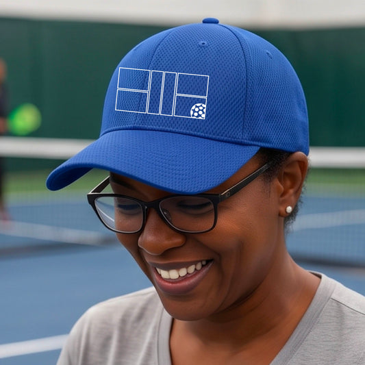 A close up of a blue baseball cap with a white pickleball court and pickleball graphic with an outdoor pickleball court.