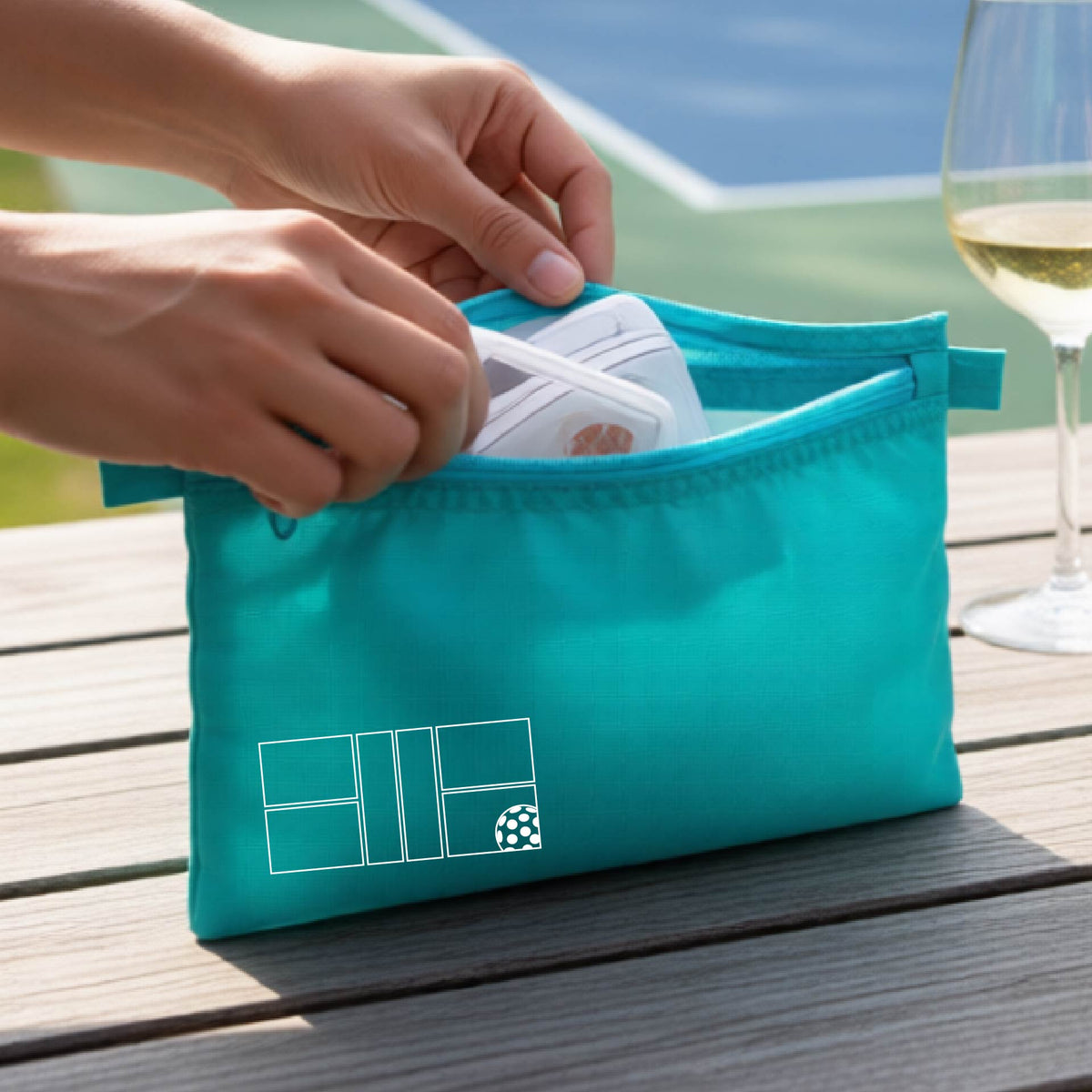 A close up of a smaller turquoise cosmetic pouch with a white pickleball court and design on the front bottom left corner. 