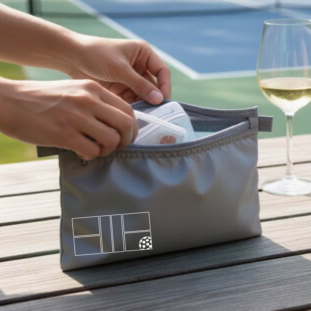 A close up of a smaller gray cosmetic pouch with a white pickleball court and design on the front bottom left corner. 