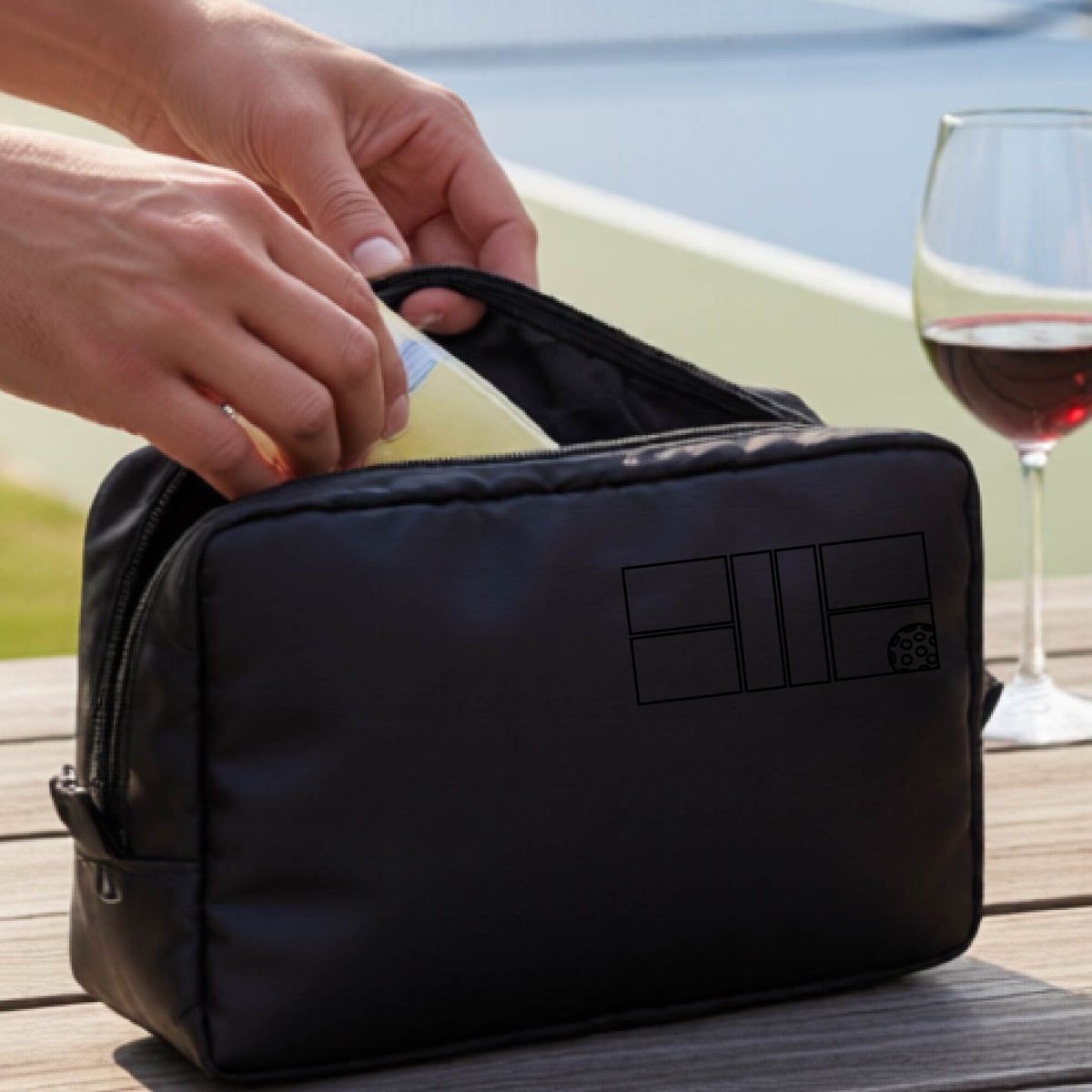A close up of a black colored pickleball cosmetic makeup bag with a black graphic of pickleball court and ball in the upper right corner. 