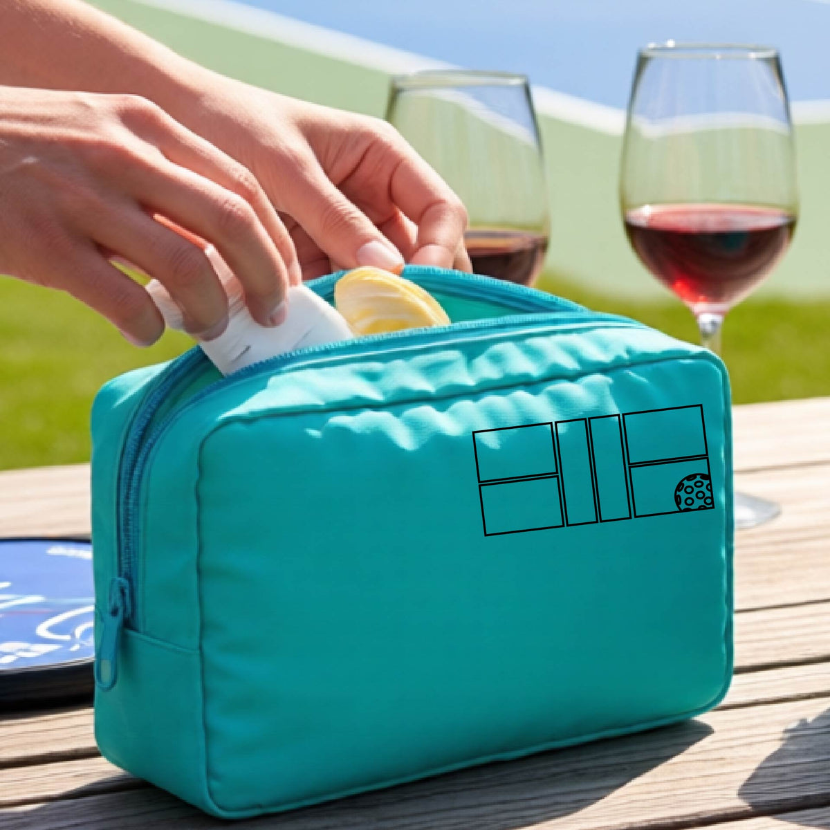 A close up of a turquoise colored pickleball cosmetic makeup bag with a black graphic of pickleball court and ball in the upper right corner. 