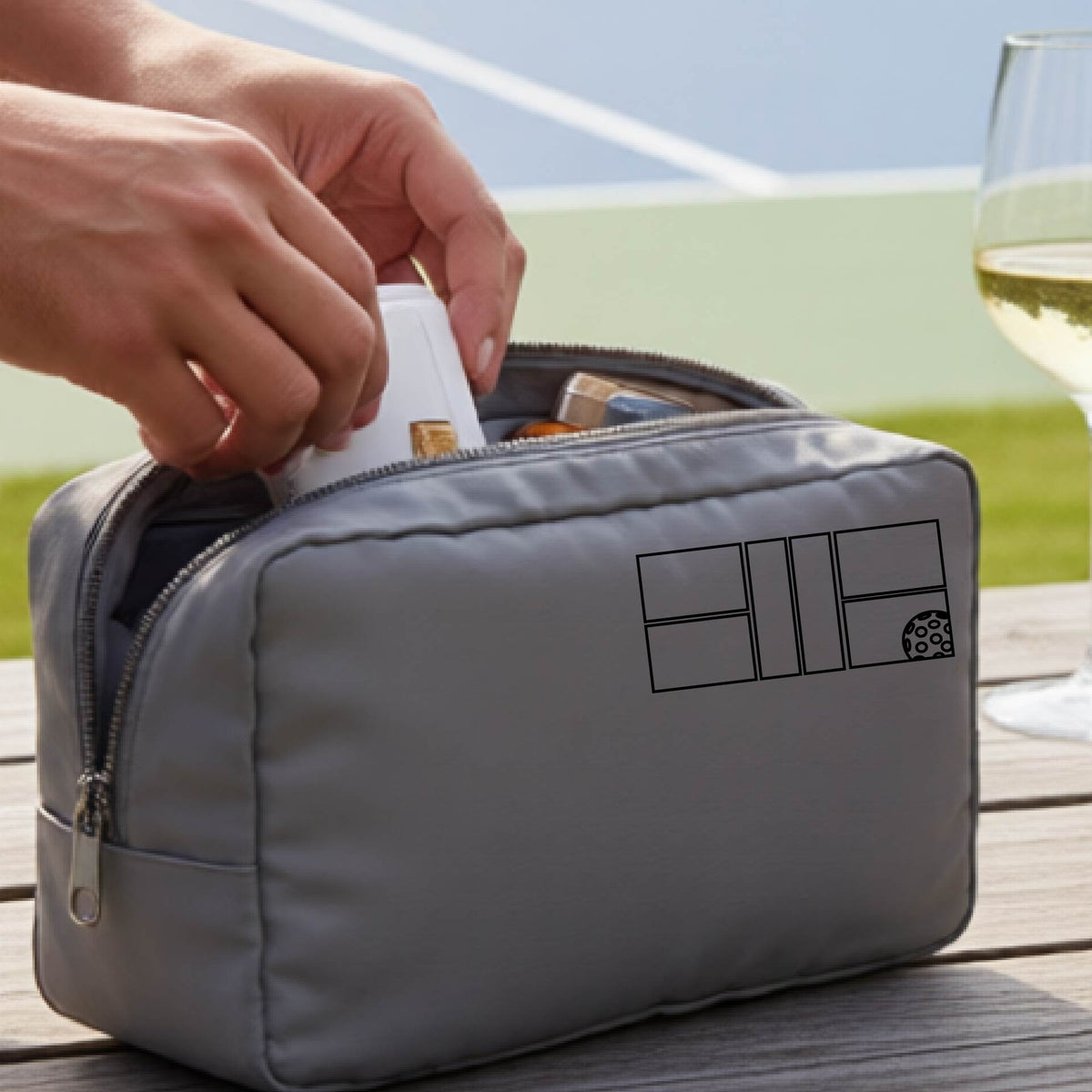 A close up of a gray colored pickleball cosmetic makeup bag with a black graphic of pickleball court and ball in the upper right corner. 