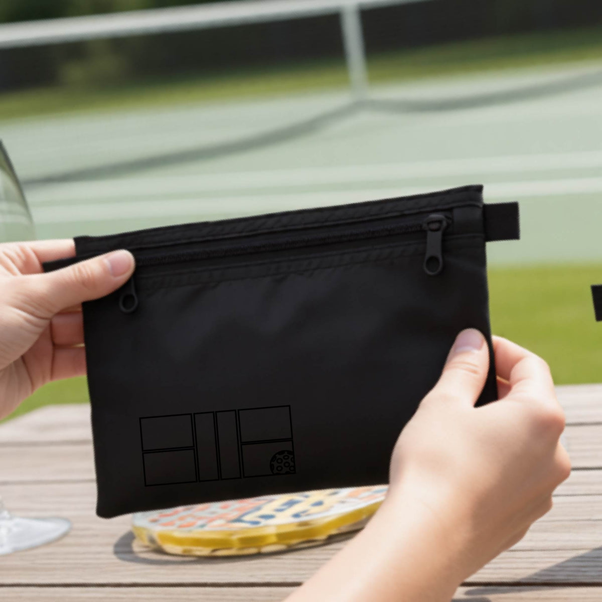 A close up of a black pickleball cosmetic makeup bag with a black graphic of pickleball court and ball in the lower left corner. 