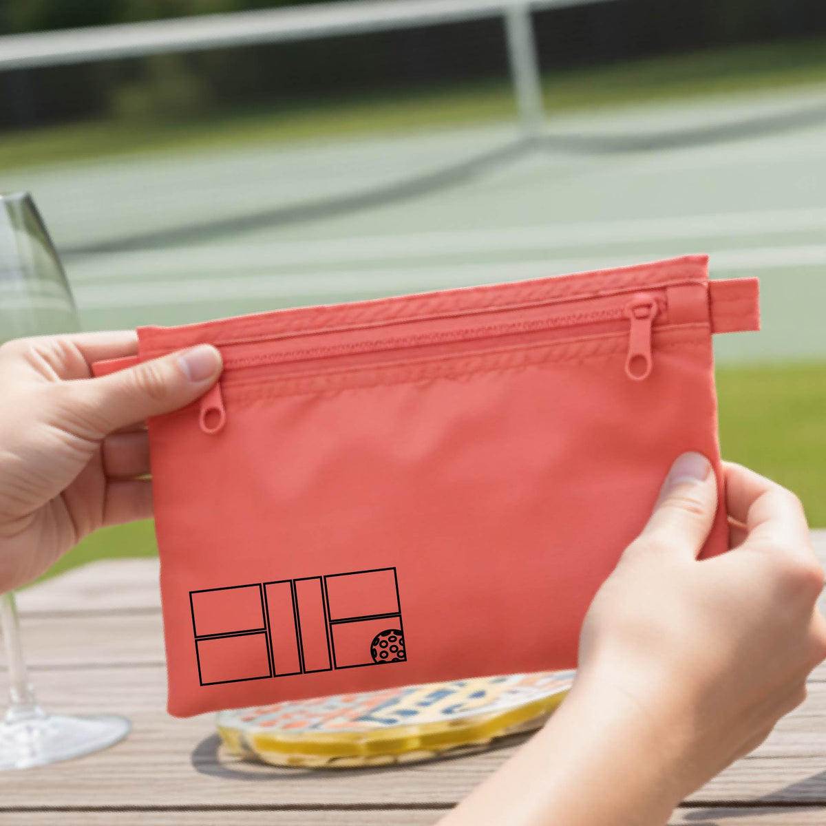 A close up of a coral colored pickleball cosmetic makeup bag with a black graphic of pickleball court and ball in the lower left corner. 