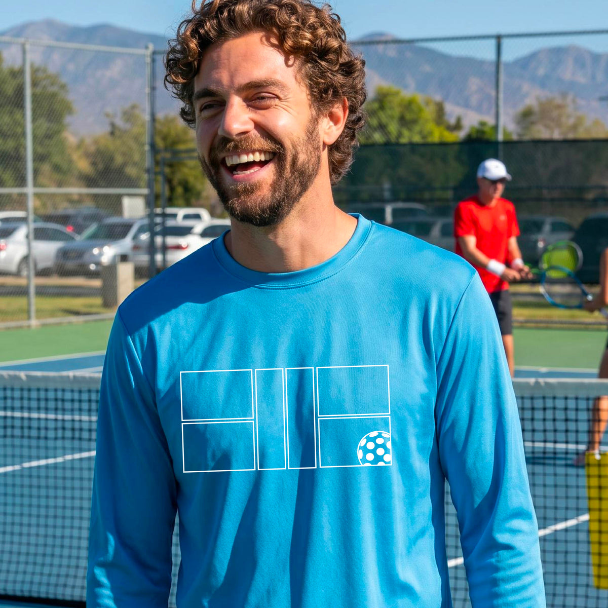 A smiling man wearing a blue long-sleeve athletic shirt with a white geometric pickleball court and ball graphic on the chest,.
