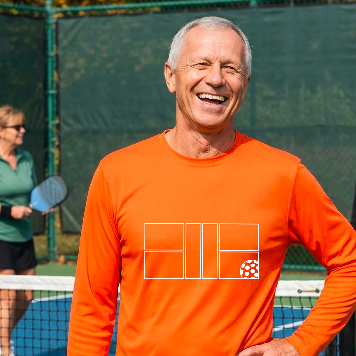 A smiling man wearing an orange long-sleeve athletic shirt with a white geometric pickleball court and ball graphic on the chest,.